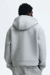 Campera con capucha y cierre frontal, color gris melange, de tejido efecto neopreno. Presenta un corte holgado (boxy) con hombros caídos y bolsillos delanteros abiertos.