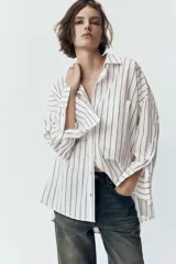 Camisa blanca con rayas verticales grises, de corte holgado, cuello solapa y mangas largas con puños abotonados. Tiene un bolsillo de parche en el pecho, pinzas en la espalda y bajo asimétrico con aberturas laterales.