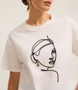 Remera blanca de algodón con cuello redondo y bordado de una cara de mujer con caravana dorada.
