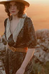 Vestido midi con estampado floral en tonos marrones y azules, cuello en V con detalle de puntilla blanca, mangas cortas abullonadas y cinturón marrón con hebilla dorada.