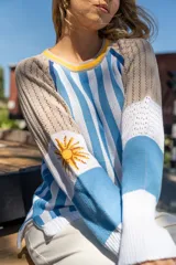 Sweater tejido a rayas verticales celestes y blancas, con cuello redondo amarillo y detalle bordado del sol de la bandera uruguaya en la manga.