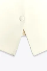 Top tipo chaleco color crema con escote recto y tirantes finos. Tiene cierre frontal con botones a tono y bajo asimétrico con abertura.