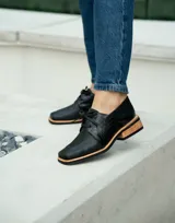 Zapato acordonado de cuero vacuno color marrón oscuro, con suela de goma, horma cuadrada y tacón bajo.