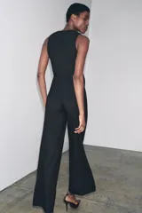 Mono largo negro de crepe, sin mangas, con escote en V y detalle superpuesto tipo chaleco cruzado con un botón. Presenta pierna ancha y cierre frontal.