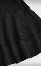 Vestido corto negro, sin mangas y con escote recto (strapless). La falda tiene un diseño con volados superpuestos.