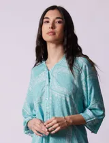 Blusa celeste con estampado de cachemir en blanco, cuello a la base, escote en V y abertura central con botones. Manga 3/4 regulable con presilla y botón, y terminación de ruedo asimétrico.