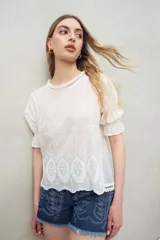 Blusa azul grisáceo de algodón con bordados frontales y terminaciones festoneadas. Tiene mangas cortas con frunces y cuello redondo con detalle de puntilla.