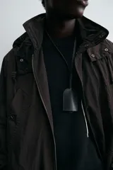 Trench relaxed fit negro confeccionado en tejido técnico ligeramente acolchado, con cuello subido y capucha desmontable. Tiene manga larga, bolsillos de vivo en pecho y de solapa en cadera, y abertura en el centro de la espalda. El cierre frontal es con cremallera oculta por solapa con botones a presión.