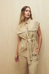 Chaleco color beige con cuello camisero, sin mangas, cierre frontal con botones y cinturón de lazo. Presenta bolsillos de parche en el pecho y pinzas verticales en el frente.
