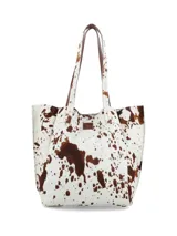 Bolso tote de cuero con pelo, estampado de vaca en blanco y marrón. Tiene asas de cuero marrón y un pequeño parche de cuero marrón con logo grabado en el frente.