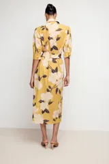 Vestido midi camisero color amarillo con estampado de flores blancas y hojas marrones. Tiene cuello solapa, mangas 3/4 con puños, cinturón de lazo ajustable y cierre frontal con botones.