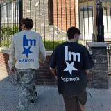 Camiseta negra con efecto acid wash, corte oversize y estampado trasero blanco con el texto "lm" sobre una estrella y el texto "lookmom" debajo.
