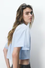 Camisa celeste de popelín con cuello solapa, escote pico y manga corta. Presenta dos bolsillos delanteros de parche y cierre frontal con botones.