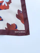 Pañuelo cuadrado de seda color crema con estampado de flores rojas, azules y blancas. Tiene borde marrón.