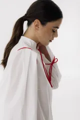 Camisa blanca de corte cropped con cuello mao y mangas largas abullonadas con lazos rojos en los puños. Presenta detalles de ribetes rojos en los hombros y lazos rojos en el cuello.
