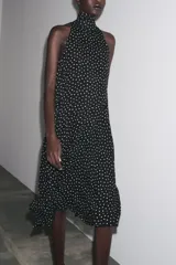 Vestido midi negro con estampado de lunares blancos, cuello halter con pañuelo integrado, sin mangas y bajo asimétrico. Cierre con abertura y botón en la nuca.