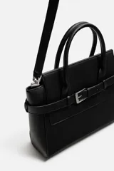 Bolso bandolera negro con detalle de cinturón, compartimento principal con cierre mediante doble imán, bolsillo interior con cierre mediante cremallera, dos asas de mano y un asa de hombro ajustable y extraíble.