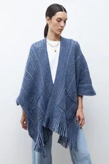 Ruana de tejido de punto en tonos beige y blanco, con patrón de espiga. Presenta cuello en V, mangas amplias, dos bolsillos frontales y flecos en el dobladillo.