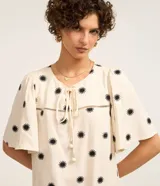 Vestido corto evasé color crema con estampado de soles negros, cuello redondo con lazo, mangas cortas y bajo con volantes.