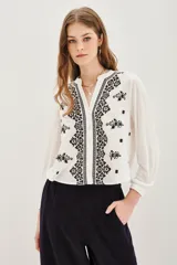 Camisa blanca con cuello mao y escote en V, con bordados negros y dorados en el frente y mangas largas con puños abotonados.