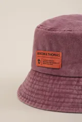 Gorro tipo bucket color lila con etiqueta naranja con el logo de la marca Benson & Thomas.