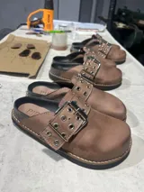 Zuecos de cuero color marrón oscuro con hebilla metálica plateada y detalle de ojales metálicos.