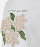 Remera blanca de algodón, con cuello redondo y manga corta. Presenta un estampado frontal con la frase "SOY FLOR. SOY LATINA" y un diseño floral con flores en relieve y hojas bordadas.
