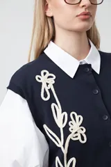 Chaleco de punto color blanco con bordado aplicado de cordón marrón en diseño floral. Presenta cierre frontal de botones al tono, cuello redondo y terminaciones acanaladas en sisas y ruedo.