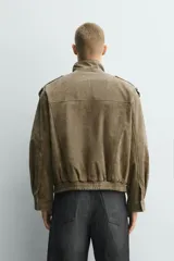 Cazadora relaxed fit de piel con acabado ante color verde militar. Tiene cuello subido, manga larga con puño abotonado, trabillas en los hombros, bolsillos de plastrón con solapa en el pecho y bolsillos de vivo en la cadera. El bajo es elástico y el cierre frontal es de cremallera oculta con solapa y botones a presión.