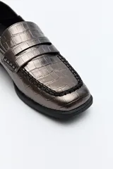 Mocasines color peltre con textura símil cuero de cocodrilo, punta cuadrada, detalle de bordado a tono en la capellada y taco bajo.