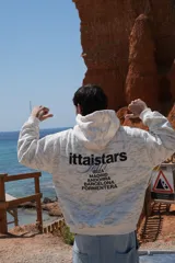 Buzo canguro con capucha, color blanco con estampado de camuflaje en tonos grises. Estampado en la espalda con el logo de la marca y nombres de ciudades.