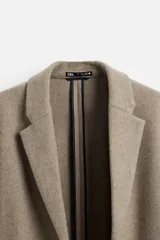 Abrigo recto color beige confeccionado en tejido con lana. Tiene cuello con solapas de muesca, manga larga con puños abotonados, bolsillos de vivo en la cadera, bolsillo interior, abertura en el centro de la espalda y cierre frontal con botonadura.