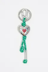 Llavero con mosquetón metálico plateado grabado con la marca Bimba y Lola. Incluye un dije central en forma de corazón metálico plateado con un corazón rojo esmaltado en el centro y letras esmaltadas alrededor. Está unido por una cuerda trenzada verde con detalles blancos.