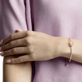 Pulsera de metal con baño en tono oro rosa, estilo brazalete abierto. Presenta un diseño central con una piedra redonda brillante suspendida dentro de una estructura ovalada. Un lado del brazalete está adornado con una hilera de cristales engastados.