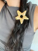 Pinza para el pelo dorada con forma de estrella. Tiene un agujero circular en el centro y una textura irregular.