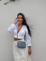 Bandolera pequeña de símil cuero color gris con textura granulada. Posee dos compartimientos principales con cierre y un bolsillo frontal con cremallera metálica. Incluye correa fina ajustable y extraíble.