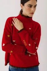 Sweater rojo de punto con cuello alto y puños acanalados en marrón. Presenta bordados de naipes en amarillo y blanco.