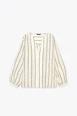 Blusa color crema con rayas verticales bordadas en negro. Tiene cuello redondo con escote en pico, mangas largas abullonadas y cierre frontal con botones.