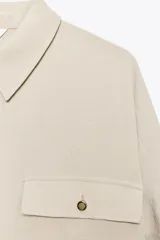 Camisa color marrón claro de cuello solapa y manga larga con trabilla y botón para remangar. Tiene un bolsillo de plastrón en el delantero, bajo asimétrico y cierre frontal con botones ocultos por solapa.