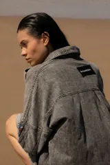 Vestido camisero de denim gris con cuello camisero, cierre frontal con botones, mangas largas con puños abotonados, bolsillos laterales y apliques bordados en el pecho y la manga. Corte holgado y largo a la rodilla.