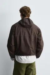 Cazadora de tejido técnico ripstop color marrón oscuro, con cuello alto y capucha ajustable, mangas largas con puños elásticos, bolsillos frontales con cremallera oculta, bajo ajustable con elástico lateral y cierre frontal con cremallera.