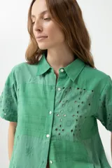 Camisa verde con cuello clásico, manga corta y cierre frontal con botones. Presenta diseño patchwork con detalles de broderie y calados.