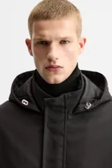 Parka negra de corte regular, confeccionada en tejido técnico acolchado. Tiene cuello alto con capucha ajustable, mangas largas con puños ajustables, bolsillos con solapa en la cadera y un bolsillo interior. El cierre frontal es con cremallera oculta por solapa con botones a presión.