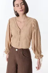 Camisa color beige con textura, escote en V, mangas largas abullonadas con lazos en los puños y cierre frontal con botones.