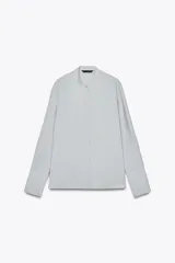 Camisa celeste fluida confeccionada en hilatura con mezcla de lyocell. Cuello redondo, manga larga con puños y bajo asimétrico con aberturas laterales. Cierre frontal con botones.