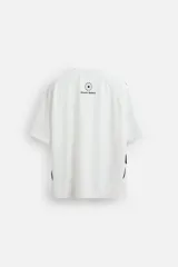Camiseta blanca de corte holgado con mangas cortas negras y cuello redondo. Presenta estampados frontales con logos y textos en azul y rojo.