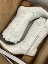 Botas texanas blancas de cuero con caña media, punta fina y cierre lateral. Presentan costuras decorativas en el empeine y el contorno de la caña.