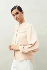 Camisa color marrón claro con cuello mao, mangas 3/4 abullonadas y cierre frontal con botones. Presenta pequeños pliegues en los hombros y detalle calado sutil.