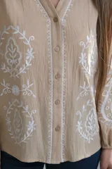 Camisa color beige con bordados florales blancos, cuello mao, mangas largas con elástico en los puños y cierre frontal con botones.