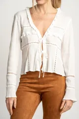 Blusa blanca de algodón con escote en V, cierre con lazos, mangas largas con puños elásticos y detalles de encaje y volados.